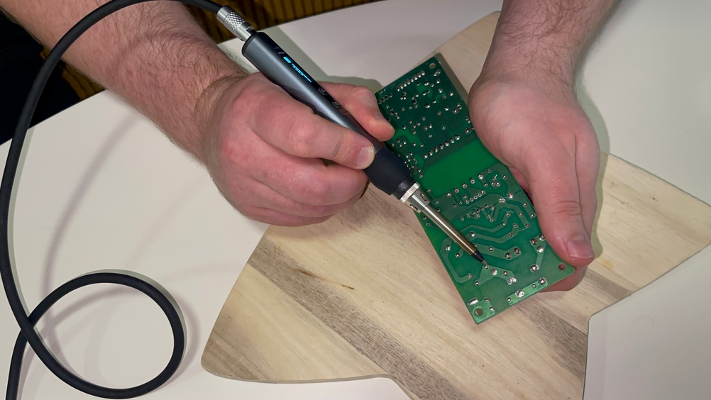 Eine Person hält eine grüne Leiterplatte in der Hand und arbeitet mit einem Kaiweets KETS02 Lötkolben an einer Lötstelle. Das Display am Lötkolben leuchtet, während die Lötspitze Kontakt zur Platine hat.
