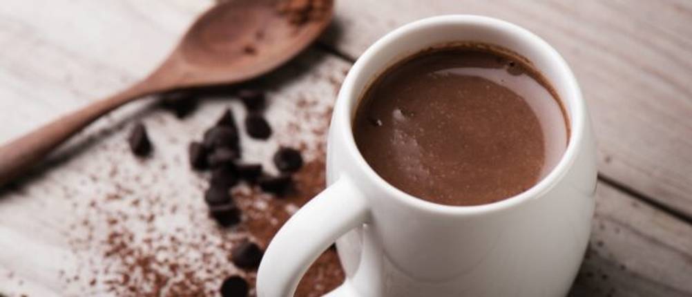Eine Tasse heiße Schokolade steht neben einem Holzlöffel und verstreutem Kakaopulver auf einem Holztisch.