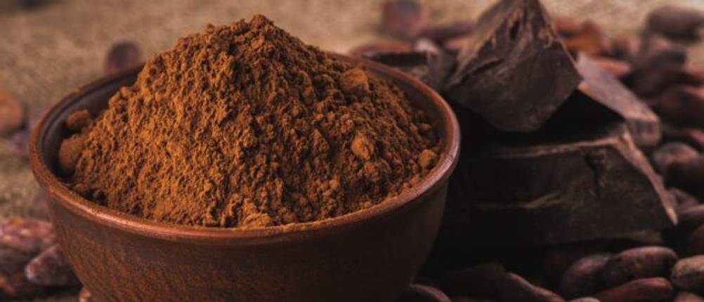 Kakaopulver in einer Schüssel auf einem Tisch, auf dem Kakaobohnen zerstreut liegen.