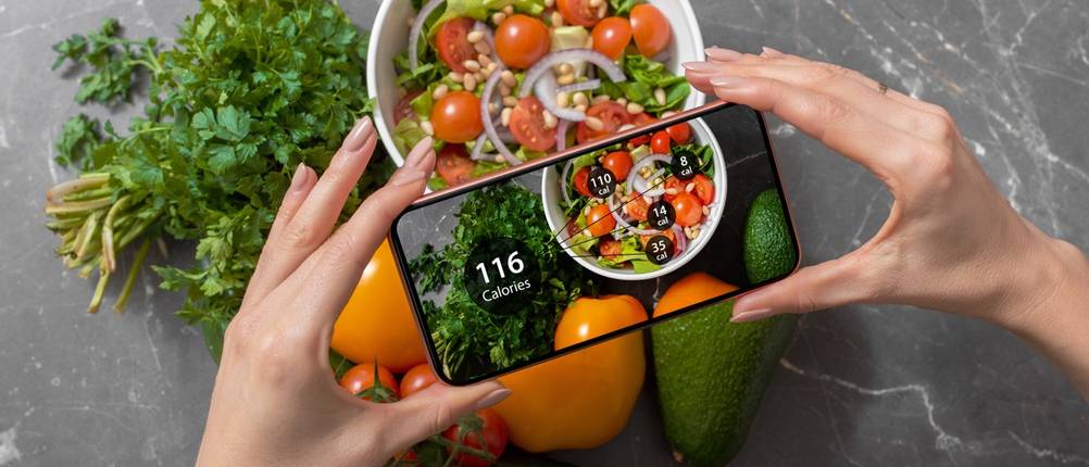 Hände halten ein Smartphone über einer Schüssel mit Salat, auf dem Display erscheint die Angabe 116 Kalorien.