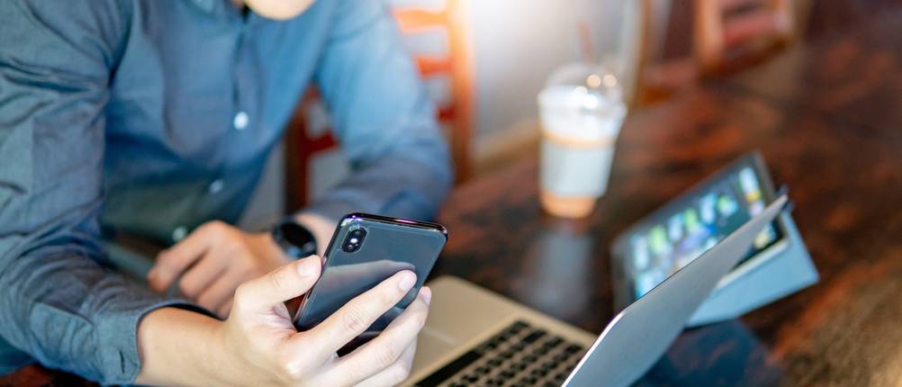 Hand mit Smartphone vor Laptop und Tablet auf einem Tisch.