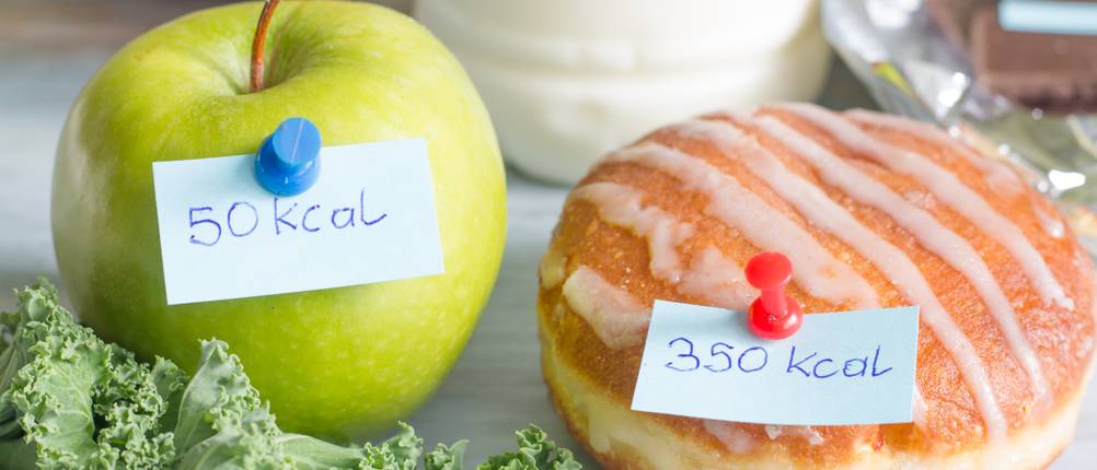 Kalorienzähler-Apps im Test: An einem Apfel und einer gebackenen Süßware hängen Zettel, auf denen die Kalorienwerte der Lebensmittel stehen.