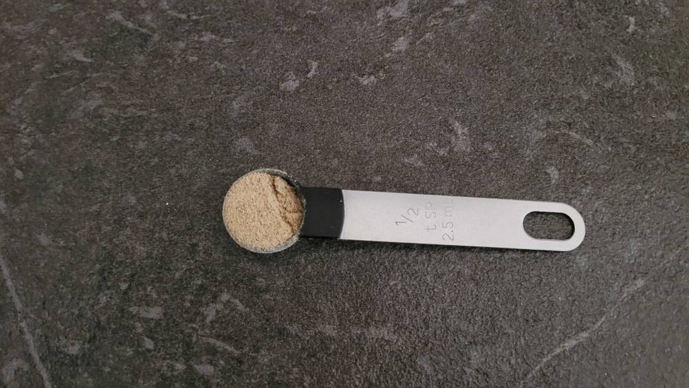 Gesamtansicht von oben auf einen Dosierlöffel mit gemahlenem weißen Pfeffer im Test, der auf einer schwarzen Oberfläche liegt.