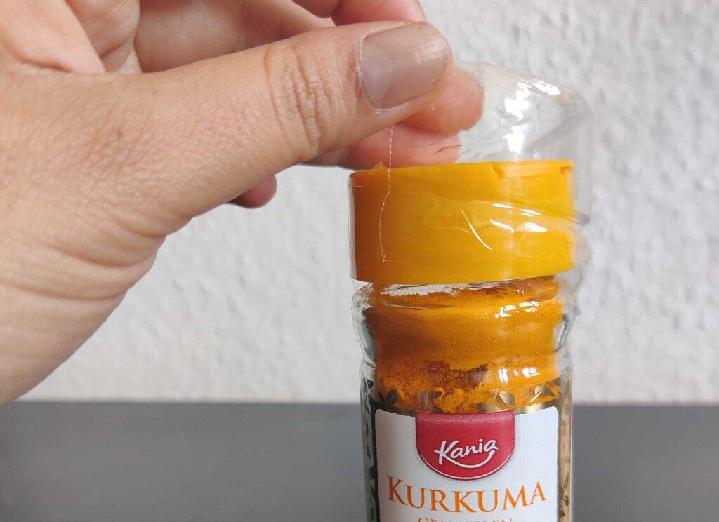 Kurkuma-Pulver im Test: Eine Hand entfernt die Vakuumfolie eines Dosier-Streuers von der Marke Kania.