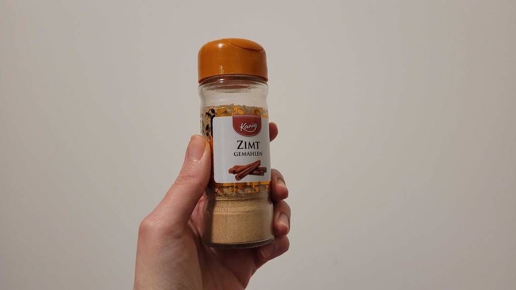 Eine Damenhand hält eine Dose getestetes Zimtpulver vor einer weißen Wand.