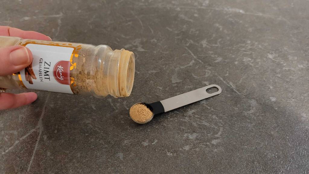 Eine Hand hält eine Dose mit getestetem Zimtpulver über einen kleinen Dosierlöffel.