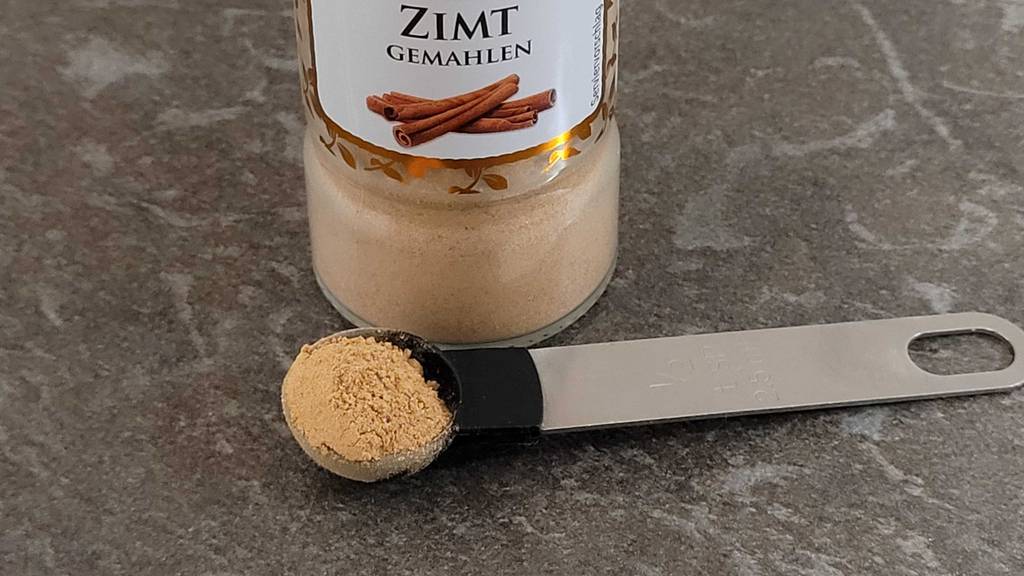 Zimtpulver im Test befindet sich auf einem Dosierlöffel vor einer transparenten Dose mit Zimt.