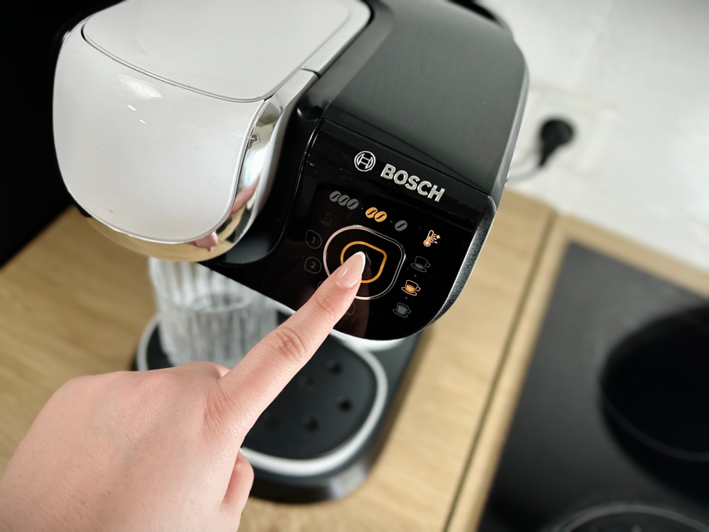 Eine Hand drückt die Starttaste an der Bosch Tassimo My Way 2. Das Touchpanel der Kapselmaschine leuchtet auf.