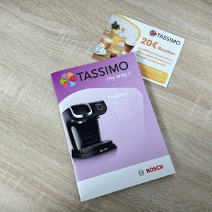 Eine Bedienungsanleitung und ein 20 €-Gutschein der Bosch Tassimo My Way 2 liegen auf einem Holztisch. Beide Dokumente sind nebeneinander platziert.
