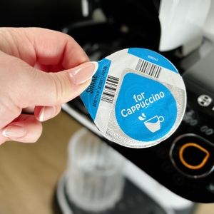 Eine Hand hält ein blaues Tassimo-Cappuccino-Pad vor die Maschine. Das Barcodeetikett auf dem Pad ist deutlich erkennbar.