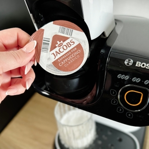 Ein Kaffeepad von Jacobs wird vor die Bosch Tassimo My Way 2 gehalten. Die Hand positioniert das Pad zum Einlegen.