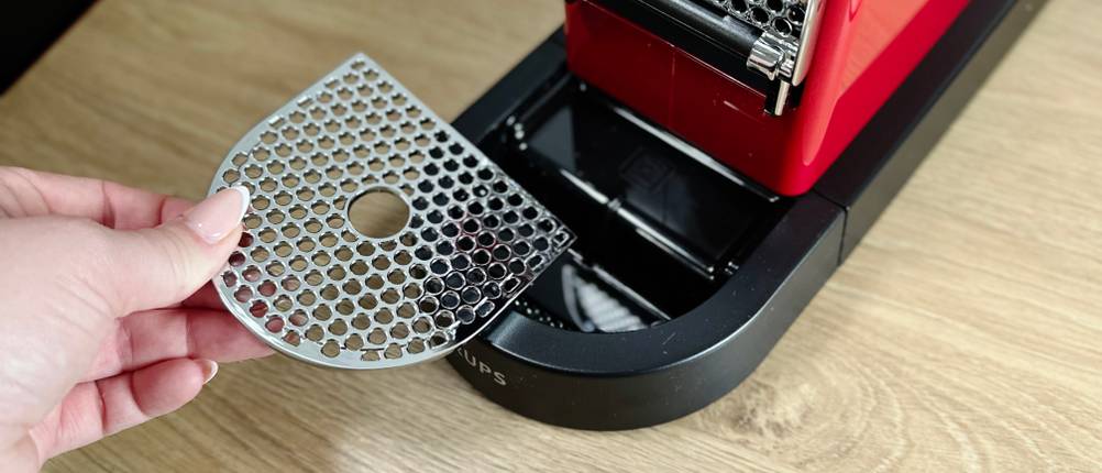 Eine Hand entnimmt das Tropfgitter der Krups Nespresso XN7415 Citiz. Das Gitter besteht aus Metall und wird zur Reinigung entfernt.