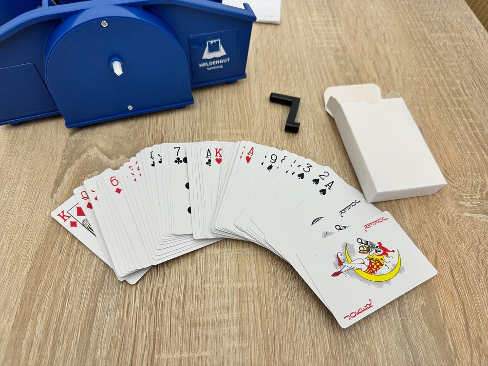 Eine Heldengut-Kartenmischmaschine liegt neben einem aufgefächerten Kartendeck und einer abgenommenen Kurbel auf einem Holztisch.