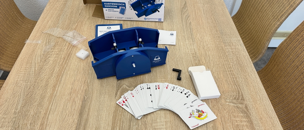 Eine Heldengut-Kartenmischmaschine liegt zusammen mit Spielkarten, Kurbel, Anleitung und Verpackung auf einem Holztisch.