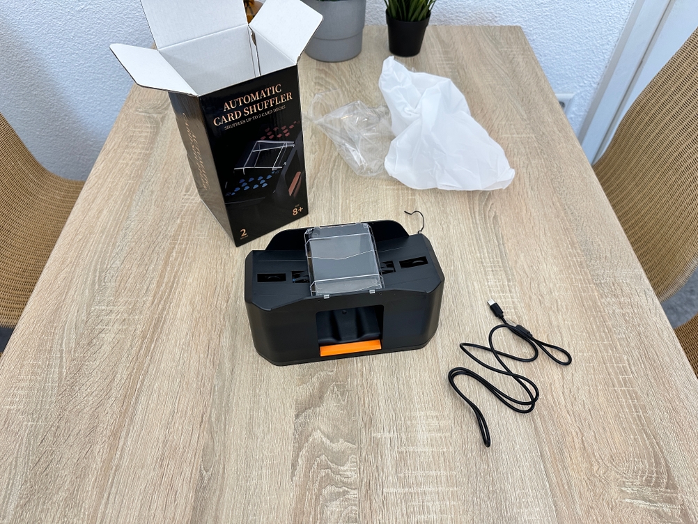 Ein Quicard-Kartenmischer JY steht vor einer geöffneten Verpackung, während ein Ladekabel daneben auf einem Tisch liegt.