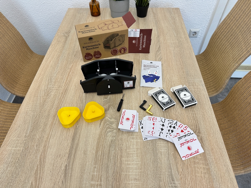Eine Weidebach-Kartenmischmaschine mit Kurbel liegt zusammen mit Spielkarten, Anleitung und Verpackung auf einem Tisch.