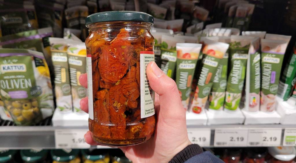 Eine Hand hält ein Glas mit getesteten getrockneten Tomaten in Öl vor einem Verkaufsregal im Supermarkt.