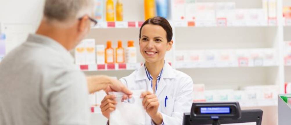 Mann kauft bei einer Apothekerin ein getestetes Produkt und bekommt es in einer weißen Tüte überreicht.