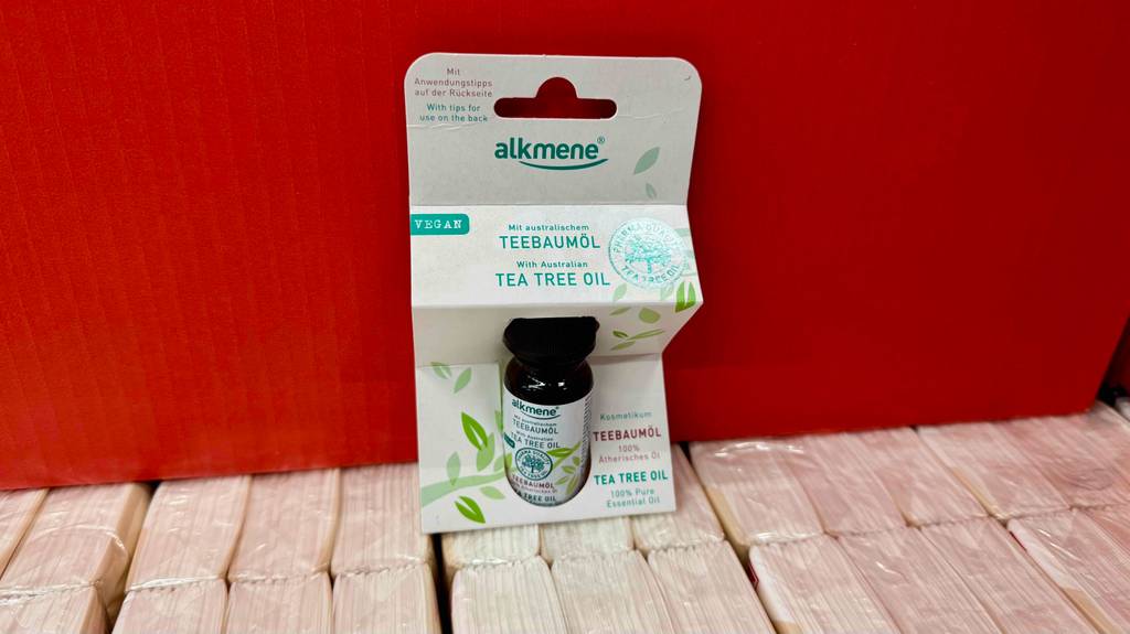 Eine Flasche mit hundertprozentig purem getestetem Alkmene-Teebaumöl steht in der Verpackung vor einer roten Wand.