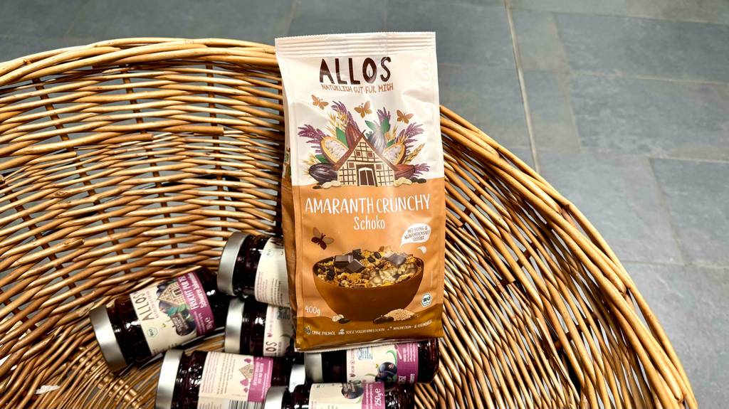 Eine 400-g-Tüte mit Allos Müsli im Test liegt in einem Korb auf Marmeladengläsern.