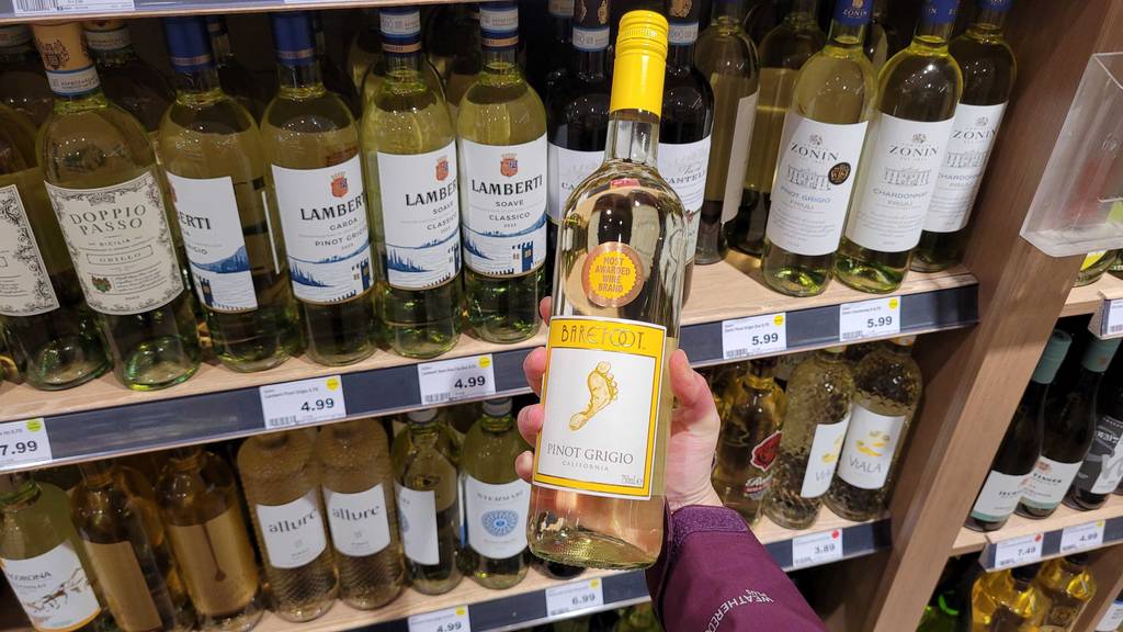 Eine Hand hält eine Flasche weißen Barefoot-Wein im Test vor einem Weinregal im Verkaufsbereich.