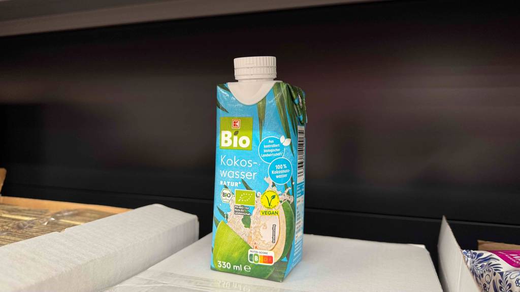 Ein Tetrapak mit weißem Schraubverschluss inklusive getestetem Bio-Kokoswasser befindet sich auf einem weißen Karton mit Lebensmitteln.