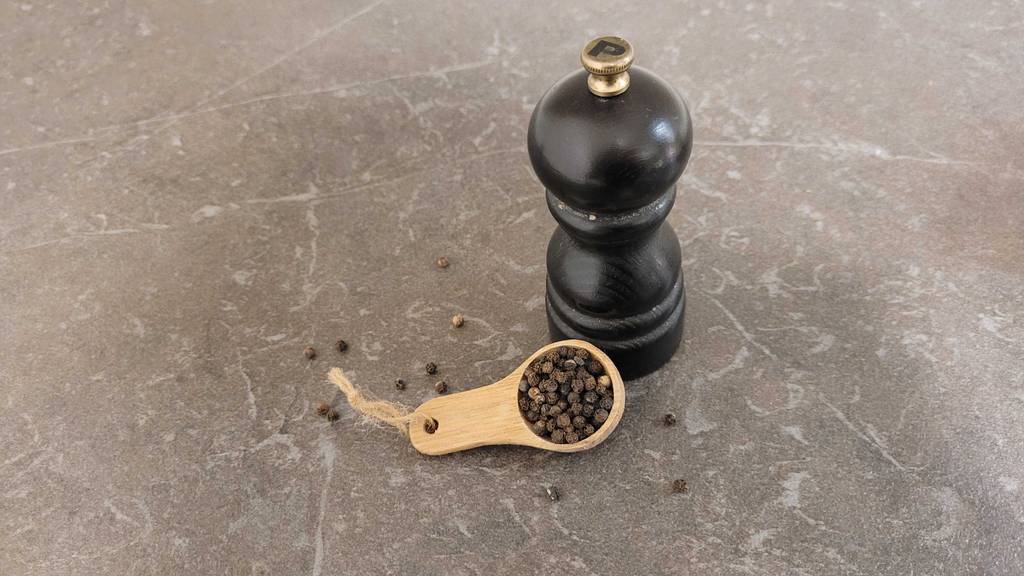 Schwarzer Pfeffer im Test liegt auf einem Holzlöffel neben einer schwarzen Pfeffermühle.