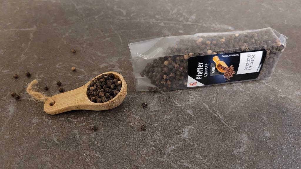 Eine Tüte mit getestetem schwarzem Pfeffer liegt neben einem Holzlöffelchen, auf dem sich schwarzer Pfeffer befindet.