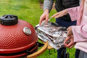 Zwei Menschen packen auf der Ablagefläche eines roten Keramikgrills etwas in Alufolie.