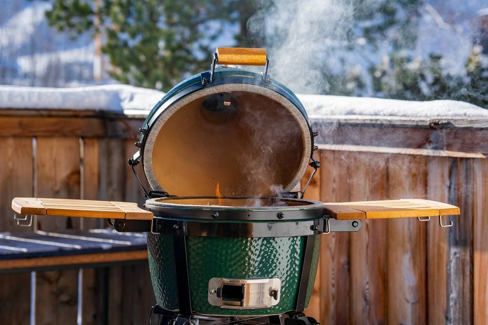 Ein offener grüner Keramikgrill steht auf einem Balkon vor einer winterlichen Landschaft.