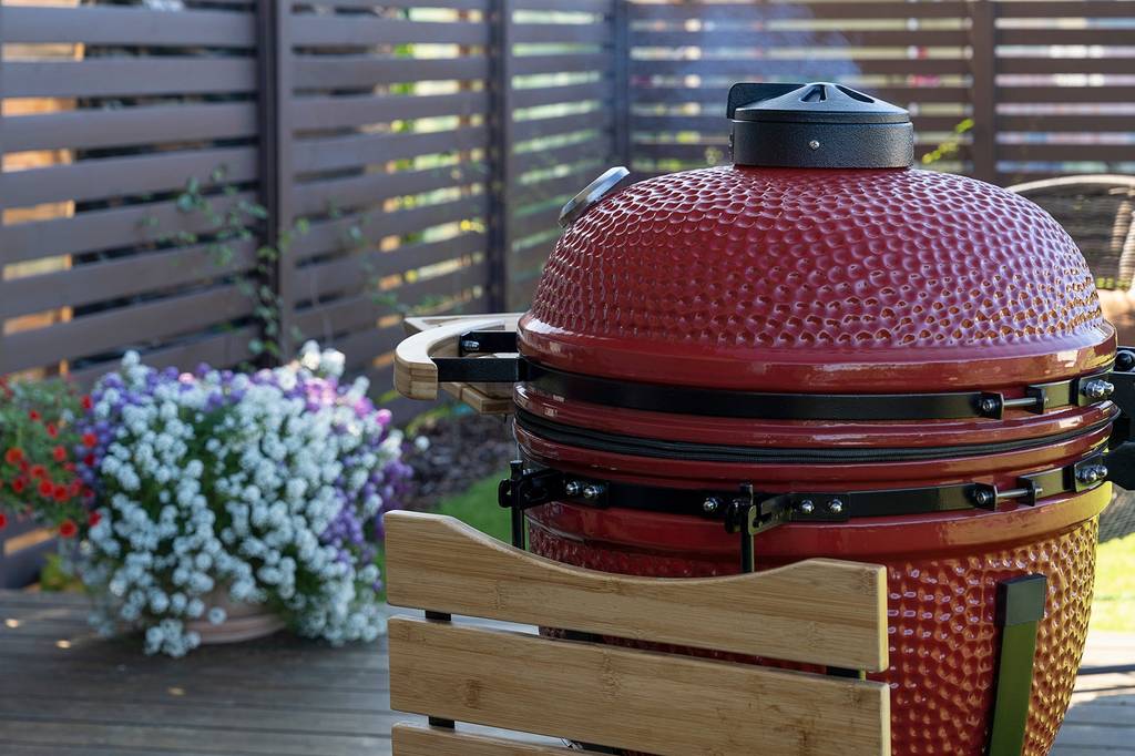 Ein geschlossener roter Keramikgrill steht in einem Garten und oben tritt leicht Rauch aus.