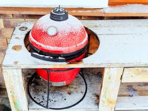 Ein roter Keramikgrill steht in einer Holzvorrichtung und ist leicht von Schnee bedeckt.