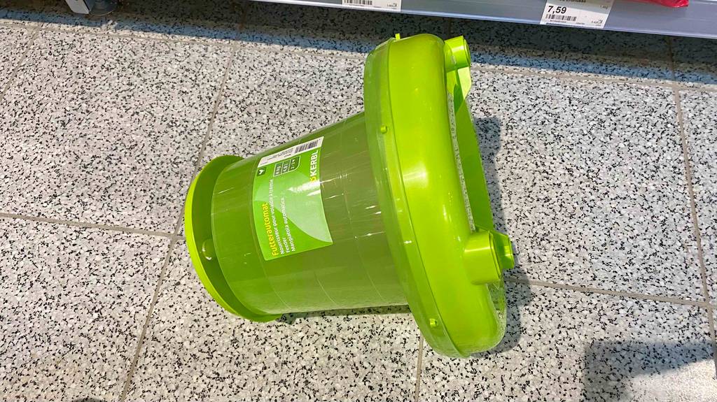 Ein getesteter Futterautomat für Hühner in giftgrüner Ausführung liegt mit leicht geöffnetem Deckel auf einem Fliesenbodenbelag.