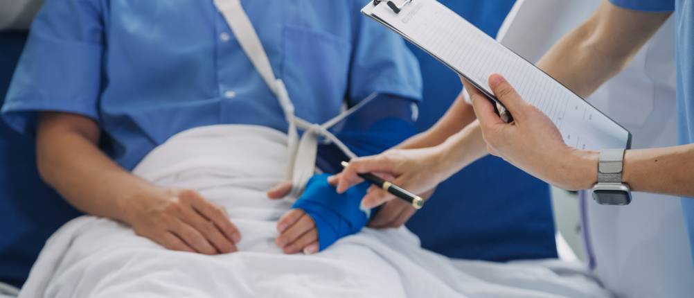 Kfz-Versicherungen im Test: Ein Mann mit Armschlaufe liegt in einem Krankenbett und wird von zwei Krankenschwestern betreut.