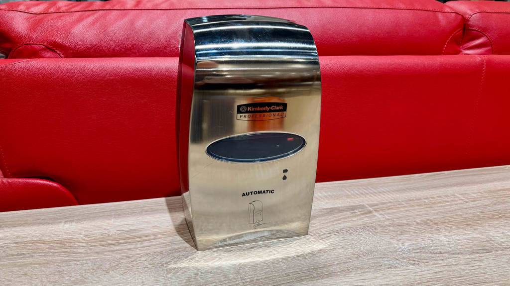 Ein Desinfektionsspender mit Sensor steht getestet auf einer Holzfläche vor einer roten Couch.