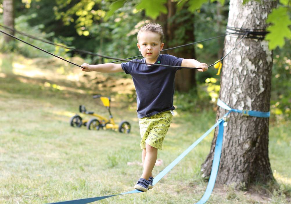 Slackline-Gestelle im Test: Ein kleiner Junge trainiert auf einer Slackline. Er hält sich an einem Sicherungsseil fest. Im Hintergrund sieht man ein Kinderfahrzeug mit vier Rädern. 