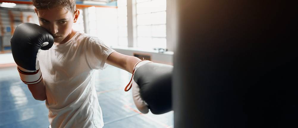 Kinder-Boxsack-Test: Ein kleiner Junge in Sportkleidung trainiert an einem schwarzen Boxsack. Er schaut grimmig.