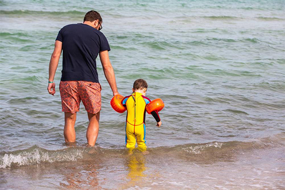 Kinder-Neoprenanzug im Test: Ein kleiner Junge in einem gelben Neoprenanzug geht mit seinem Papa ins Meer.