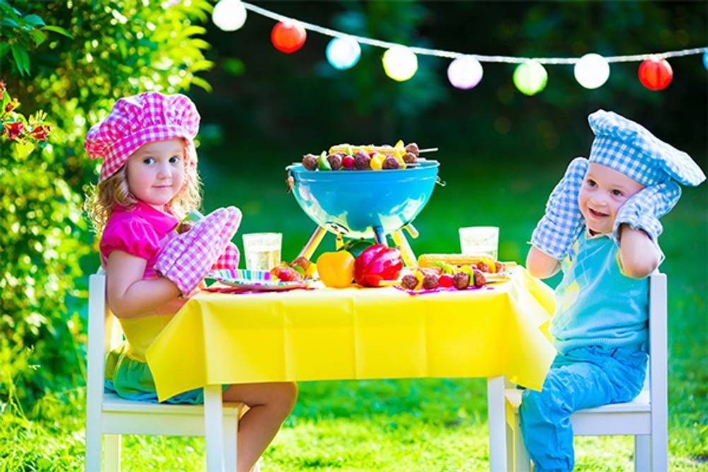 Kindergrill im Test: Zwei Kinder im Garten, die mit einem blauen Tischgrill an einem bunt gedeckten Tisch sitzen.