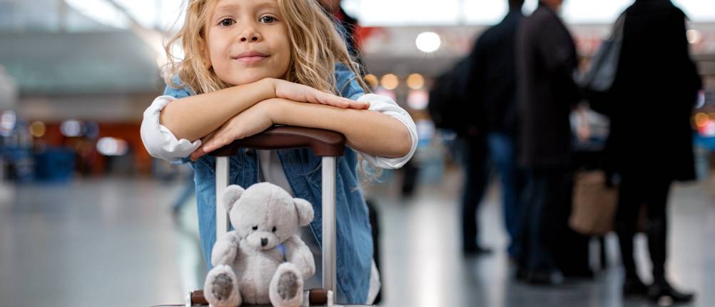 Kinderkoffer im Test: blondes Kind im Flughafenterminal, auf den Teleskopgriff eines Koffers gestützt und vor sich sein Kuscheltier