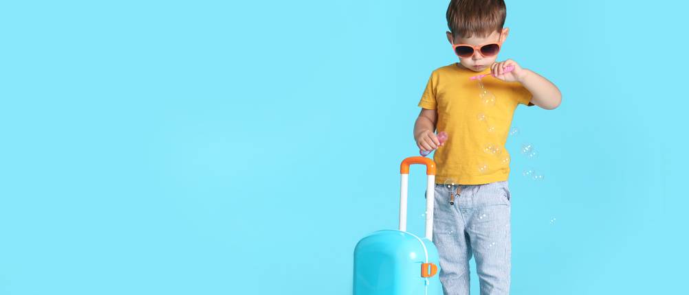 Kinderkoffer im Test: kleiner Junge mit Sonnenbrille, der Seifenblasen macht und Koffer vor blauem Hintergrund