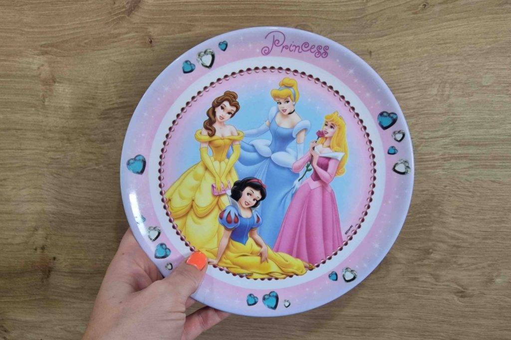 Hand hält einen getesteten Kinderteller mit Prinzessinnen-Motiv über Holzfläche.
