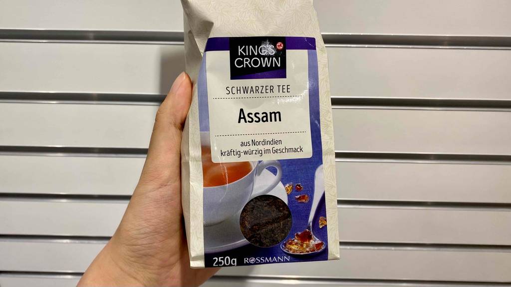 Im Test: Hand hält 250 Gramm Packung Kings Crown Assam Tee vor weißer Lamellenwand.