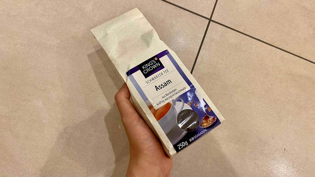 Im Test: Hand hält eine Packung Kings Crown Assam Tee schräg über hellen Fliesen.