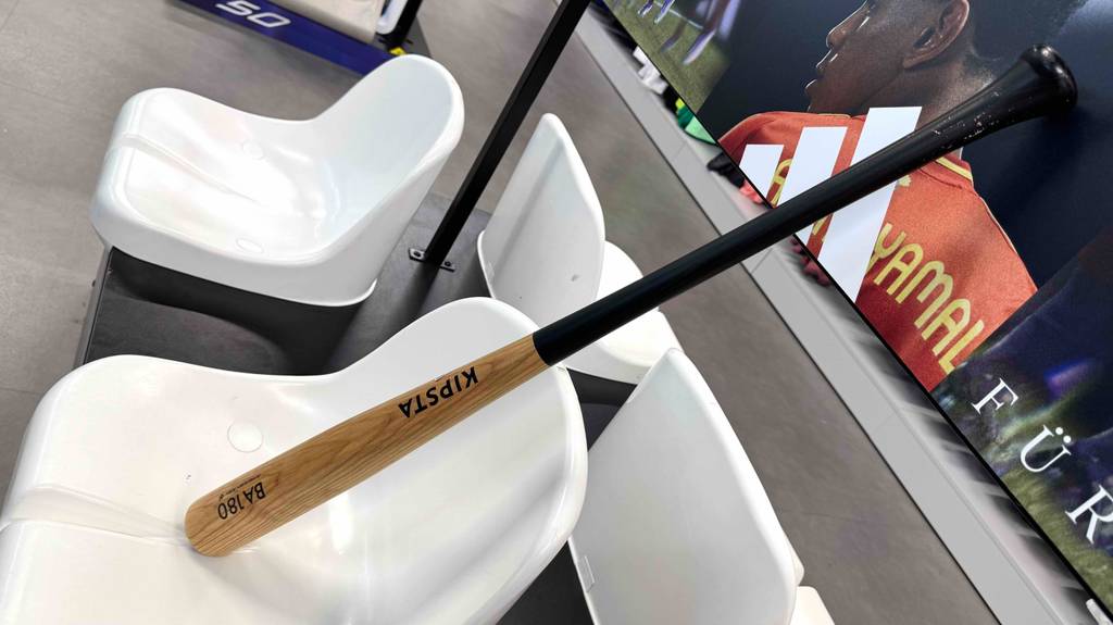 Der Baseballschläger im Test, ausgestattet mit einem Holzschlagbereich, lehnt an einem weißen Stuhl im Sportgeschäft.