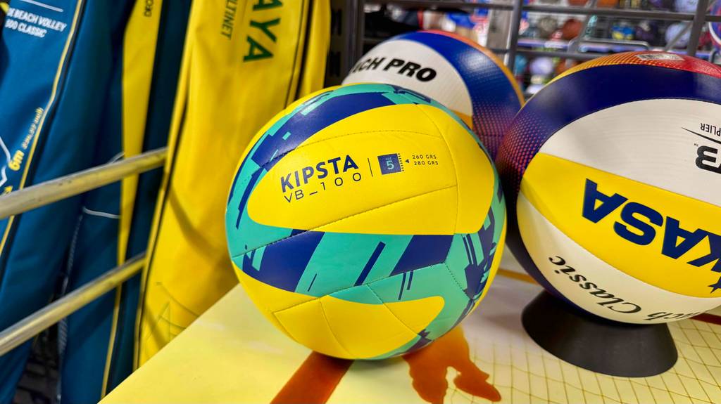 Der türkis-gelbe Volleyball im Test, Typ VB_100 von KIPSTA, liegt neben weiteren Bällen im Verkaufsregal.