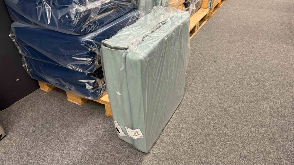 Getestet: Eine verpackte Klappmatratze steht seitlich vor einer Holzpalette mit anderen Produkten.