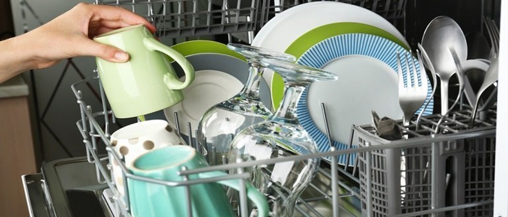 Klarspüler in einer offenen Spülmaschine mit vollem Geschirr, in die eine Frau eine grüne Tasse hineinstellt