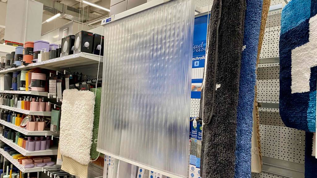 Ein semitransparentes Duschrollo im Test inklusive Seitenzug, hängt heruntergelassen in einem Verkaufsbereich neben weiteren Badezimmerartikeln, wie Duschvorleger und Seifenspendern.