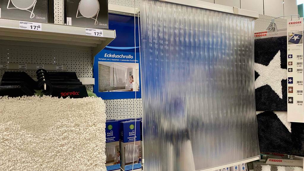 Duschrollo-Test: ein semitransparentes Duschrollo mit Seitenzug, vor einem Plakat mit Foto als Eckduschrollo, hängt in einem Verkaufsbereich neben weiteren Badezimmerartikeln.